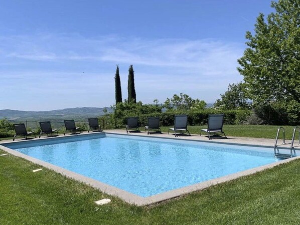 Une piscine rectangulaire à l’eau d’un bleu limpide, entourée d’une pelouse bien entretenue. Six chaises longues noires sont alignées le long du bord de la piscine, et celle-ci se trouve devant un décor composé d’arbres verdoyants et d’un ciel dégagé.