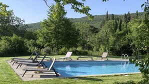 Une piscine avec des chaises longues et une pelouse, le tout entouré d’arbres et de collines.