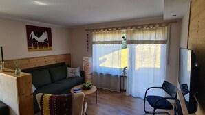 A cozy living room with a wooden floor, a green sofa, a small table with a bowl of fruit, a TV, and a window with curtains. - "Chalet Imberg", Oberstaufen-Steibis (Oberstaufen)
