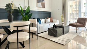 A modern living room with a white sofa, a coffee table, and a dining area with chairs and a vase of flowers. - Silkhaus Premium 1bdr in Dubai Production City (Dubai)