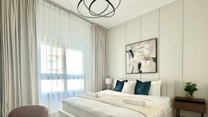 A modern bedroom with a large bed, a bedside table with a lamp, and a window with sheer curtains. - Silkhaus Premium 1bdr in Dubai Production City (Dubai)