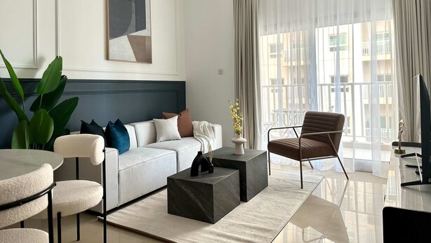 A modern living room with a white sofa, a coffee table, a brown armchair, and a large window with sheer curtains. - Silkhaus Premium 1bdr in Dubai Production City (Dubai)
