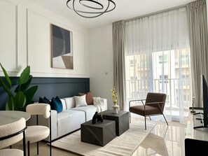 A modern living room with a white sofa, a coffee table, a brown armchair, and a large window with sheer curtains. - Silkhaus Premium 1bdr in Dubai Production City (Dubai)
