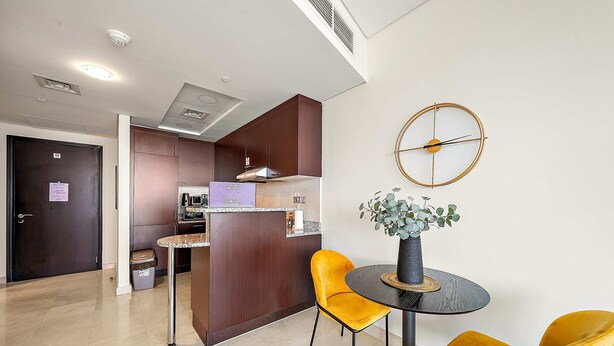 A modern kitchen with dark wooden cabinets, a round table with two yellow chairs, and a wall-mounted clock. - Silkhaus Comfy Studio in Sky Gardens Difc Views (Dubai)