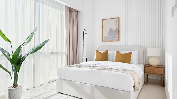 Quarto de hotel moderno com uma cama grande, uma planta em vaso, uma mesa de cabeceira com abajur e um quadro na parede.