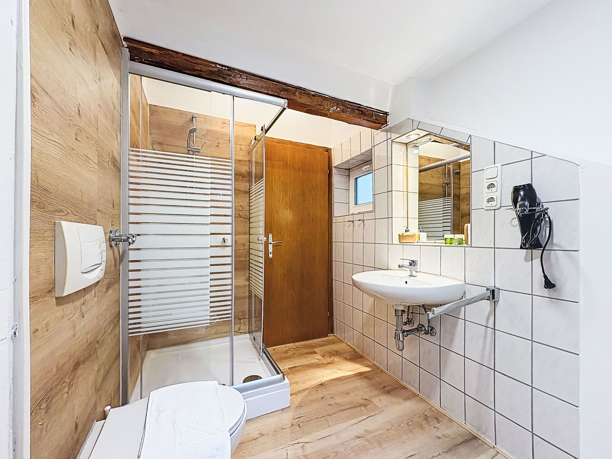 Ein modernes Badezimmer mit Duschkabine aus Glas, Holztür, weißem Waschbecken und Spiegel mit Wandhaartrockner.