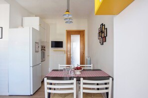 Cocina compacta con armarios blancos, un refrigerador, un microondas y un televisor pequeño. Hay una mesa de comedor con sillas y mantel a cuadros rojo y blanco.
