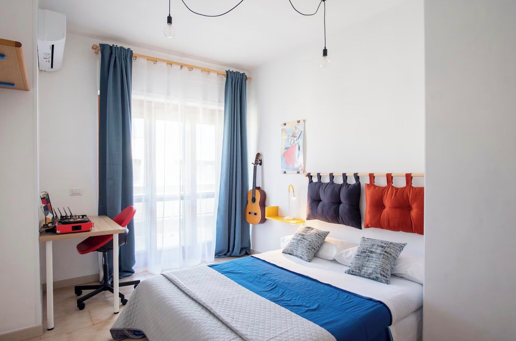 A bedroom with a bed, a desk, a chair, a guitar, and a window with curtains.