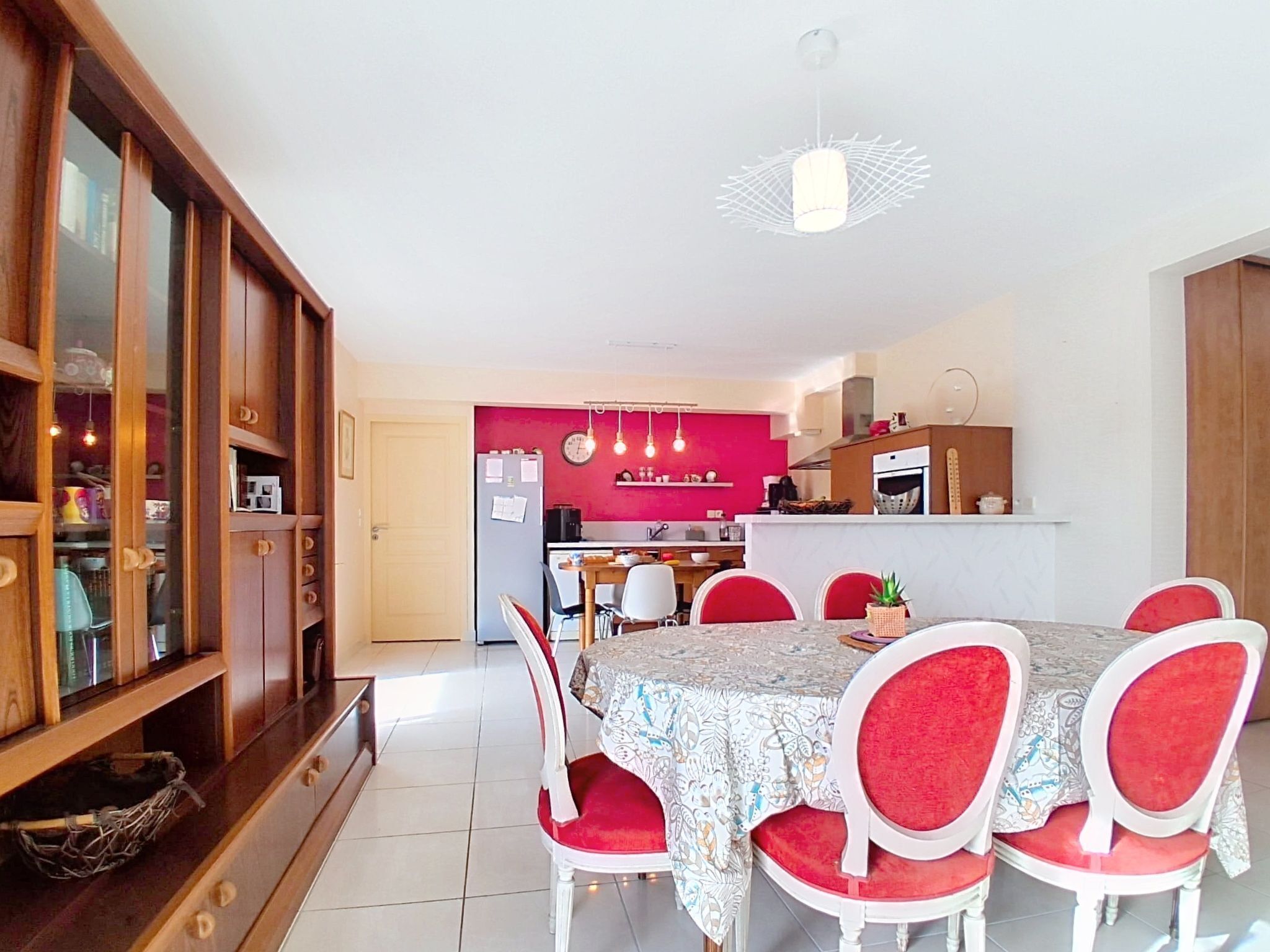 Zona de dinar amb taula preparat per a sis, envoltada de cadires vermelles tapissades. Es veu una cuina al fons amb una paret vermella d'accentuació.