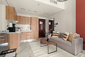 A modern kitchen with wooden cabinets, a built-in microwave, and a coffee machine. There is a grey sofa with decorative pillows and a small wooden coffee table. - Silkhaus City View 1bdr In Maryah Vista Tower (Abu Dhabi)