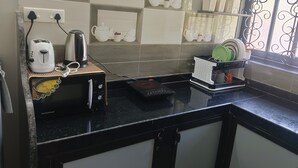 Una cocina con cubierta negra, microondas, hervidor de agua y tostadora. Hay estantes con vajilla blanca y una ventana con rejas decorativas de hierro.