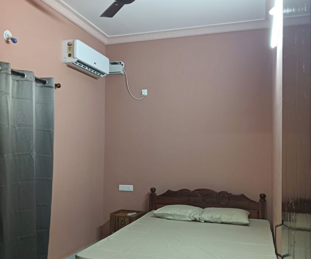 A bedroom with a bed, a wooden headboard, a nightstand, a wall-mounted air conditioner, and a curtain.