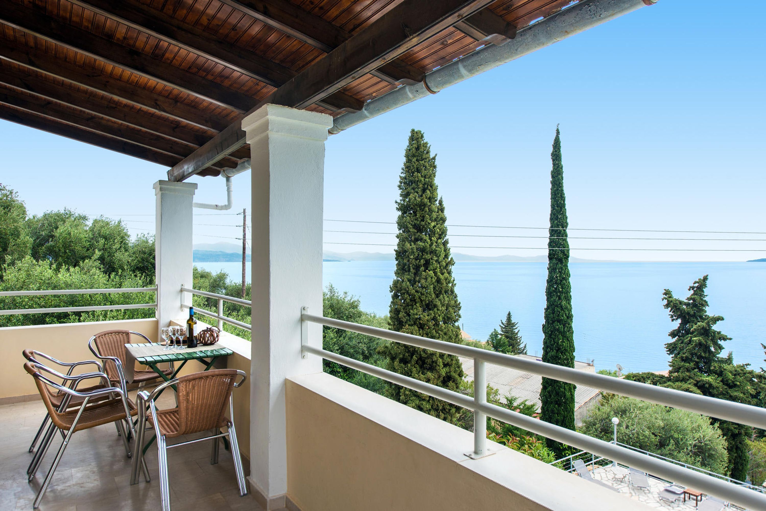A balcony with wicker chairs and a table, overlooking a scenic view of the sea and mountains.