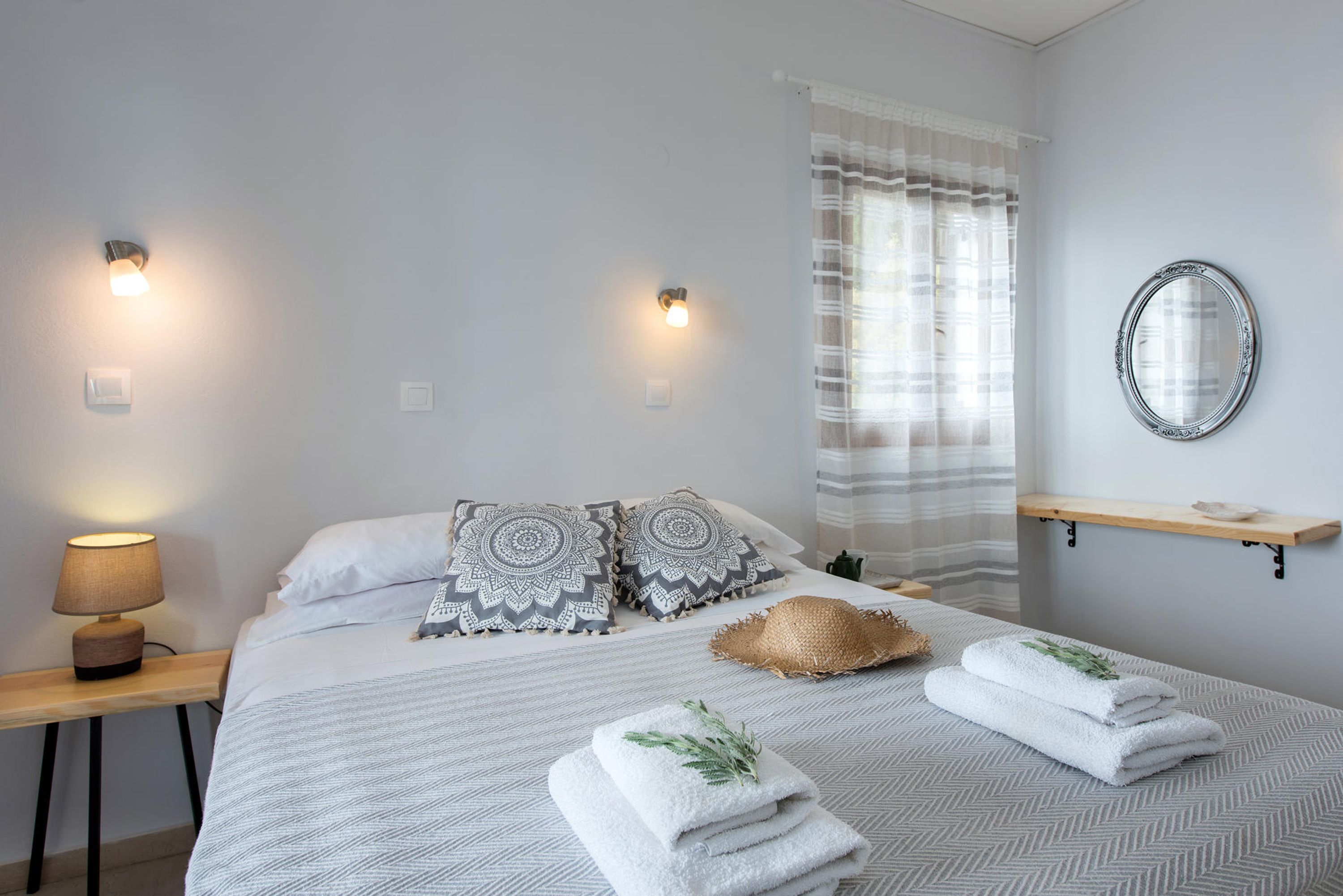 Een netjes ingerichte slaapkamer met een bed, kussens, een bedlampje, een spiegel en handdoeken op het bed.