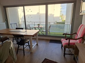 Une pièce avec une table en bois, des chaises, un fauteuil à rayures et une vue sur un balcon, visible à travers de grandes fenêtres.