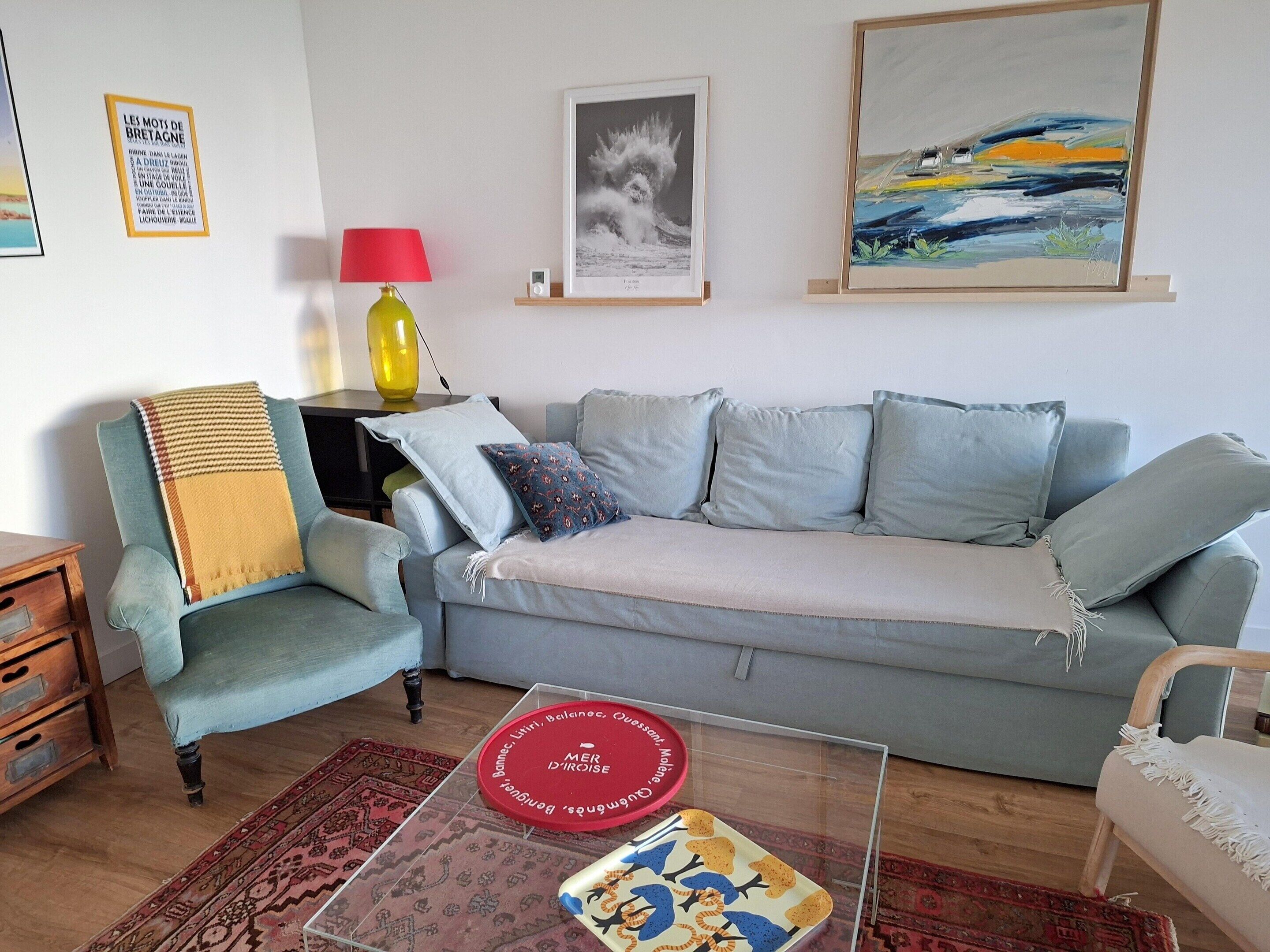 A living room with a light blue sofa, a patterned armchair, a red lamp, and a coffee table with a red and blue plate.