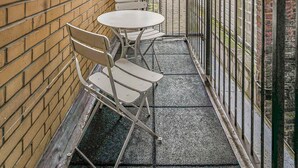 Un estrecho pasillo exterior con paredes de ladrillo, barandilla de metal y una pequeña mesa con sillas.