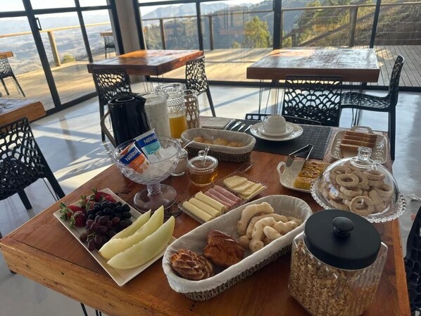 A breakfast buffet with various fruits, pastries, and condiments on a wooden table in a room with large windows. - Pousada Dona Pedra (Córrego do Bom Jesus)