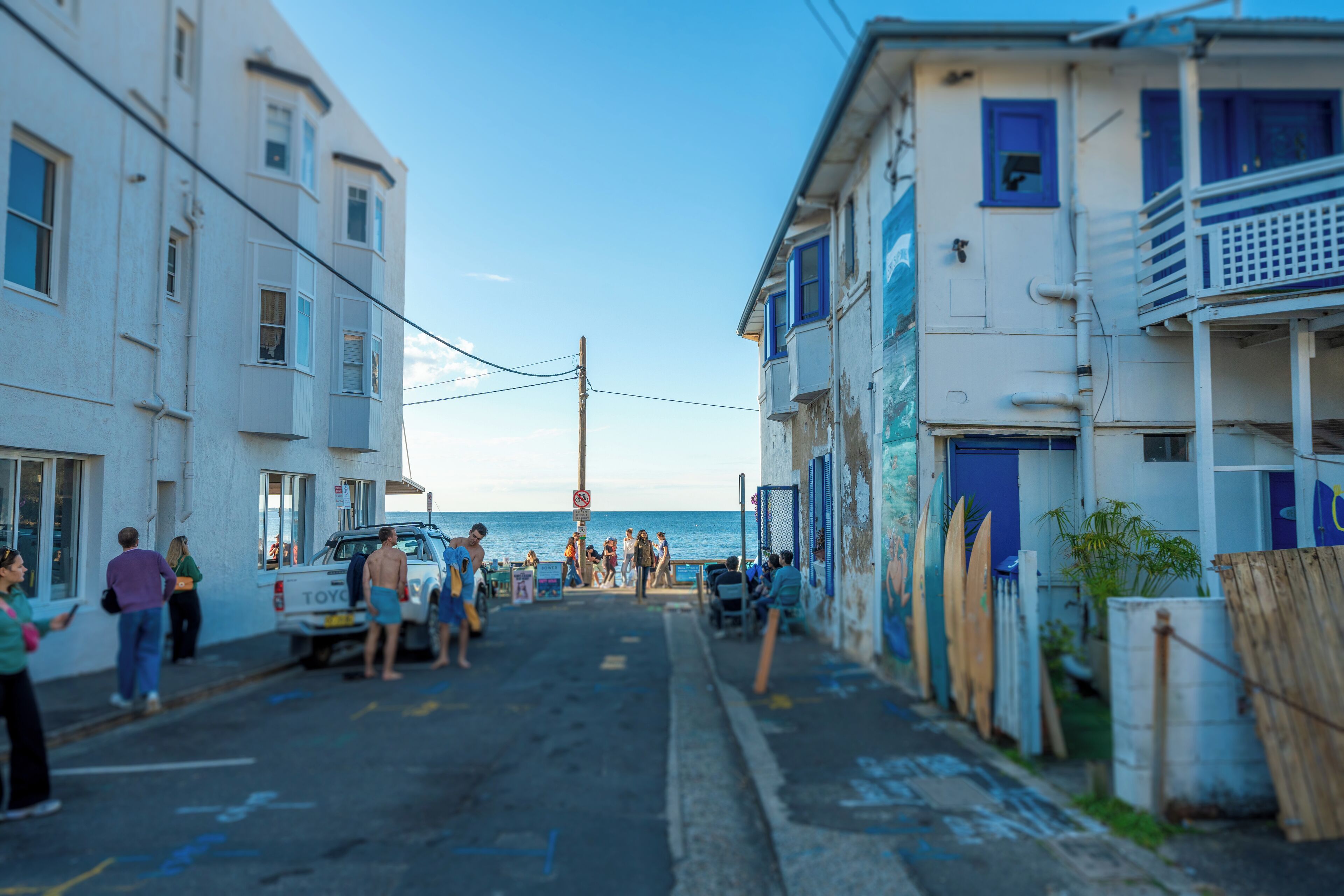 A narrow street with buildings on either side, a parked car, and people walking.