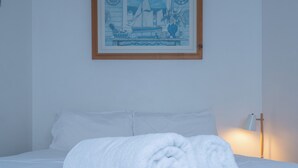 Ein ordentlich bezogenes Bett, ein aufgerolltes weißes Handtuch, ein gerahmtes Bild eines Segelschiffs an der Wand und eine Nachttischlampe.