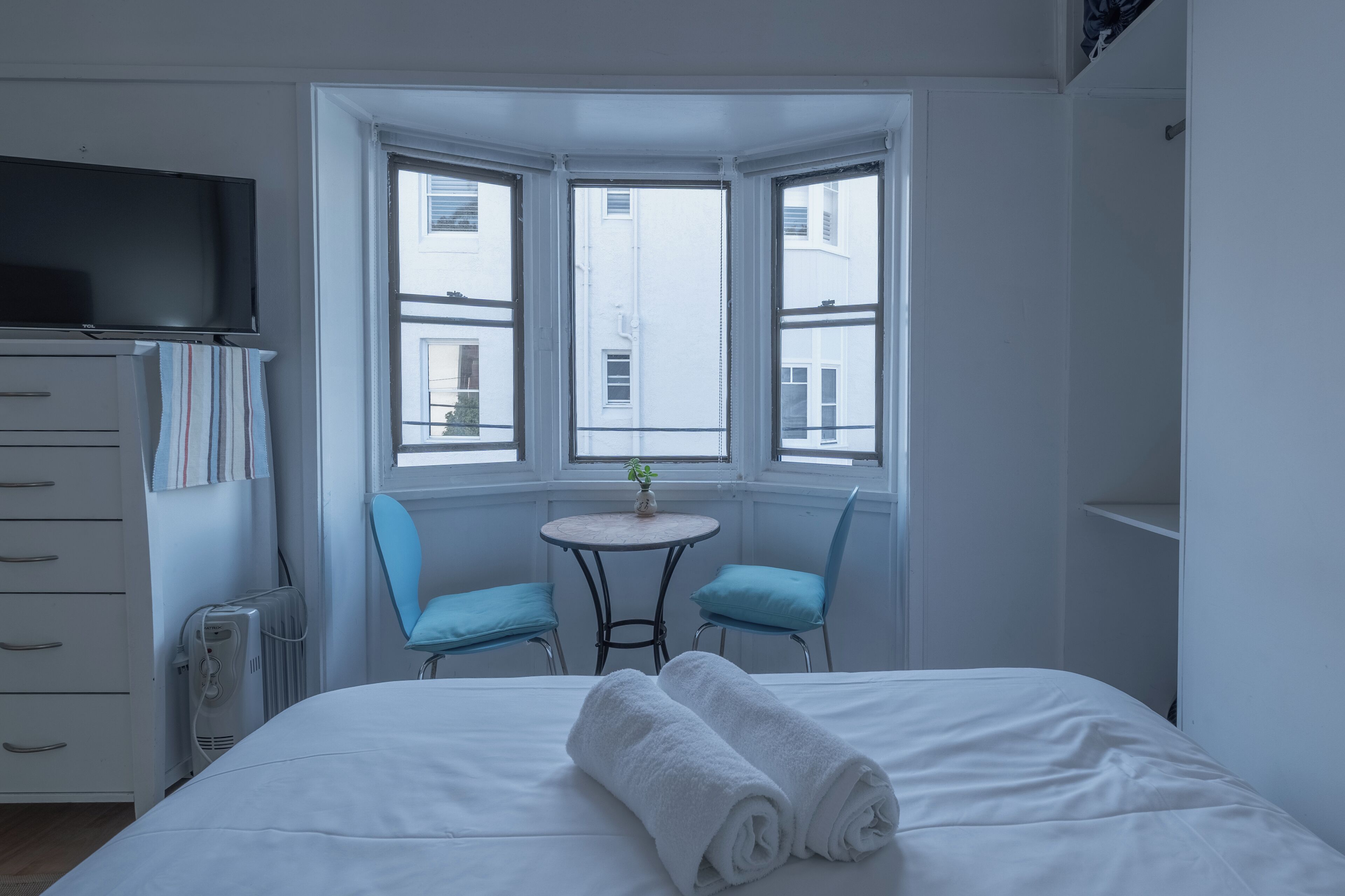 A hotel room with a bed, a TV, a radiator, a small table with chairs, and a bay window with a view of buildings.