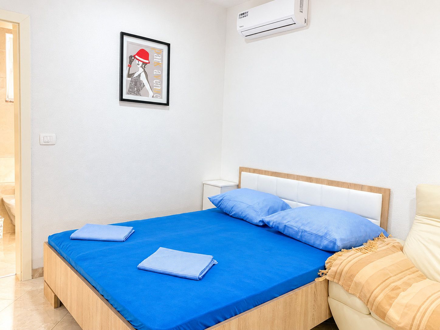 A bedroom with a bed, a framed picture on the wall, and an air conditioning unit.