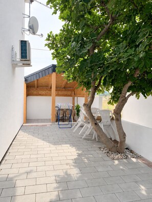 Ein überdachter Außenbereich mit Tisch, Stühlen, einem Baum und einer Satellitenschüssel an der Wand.