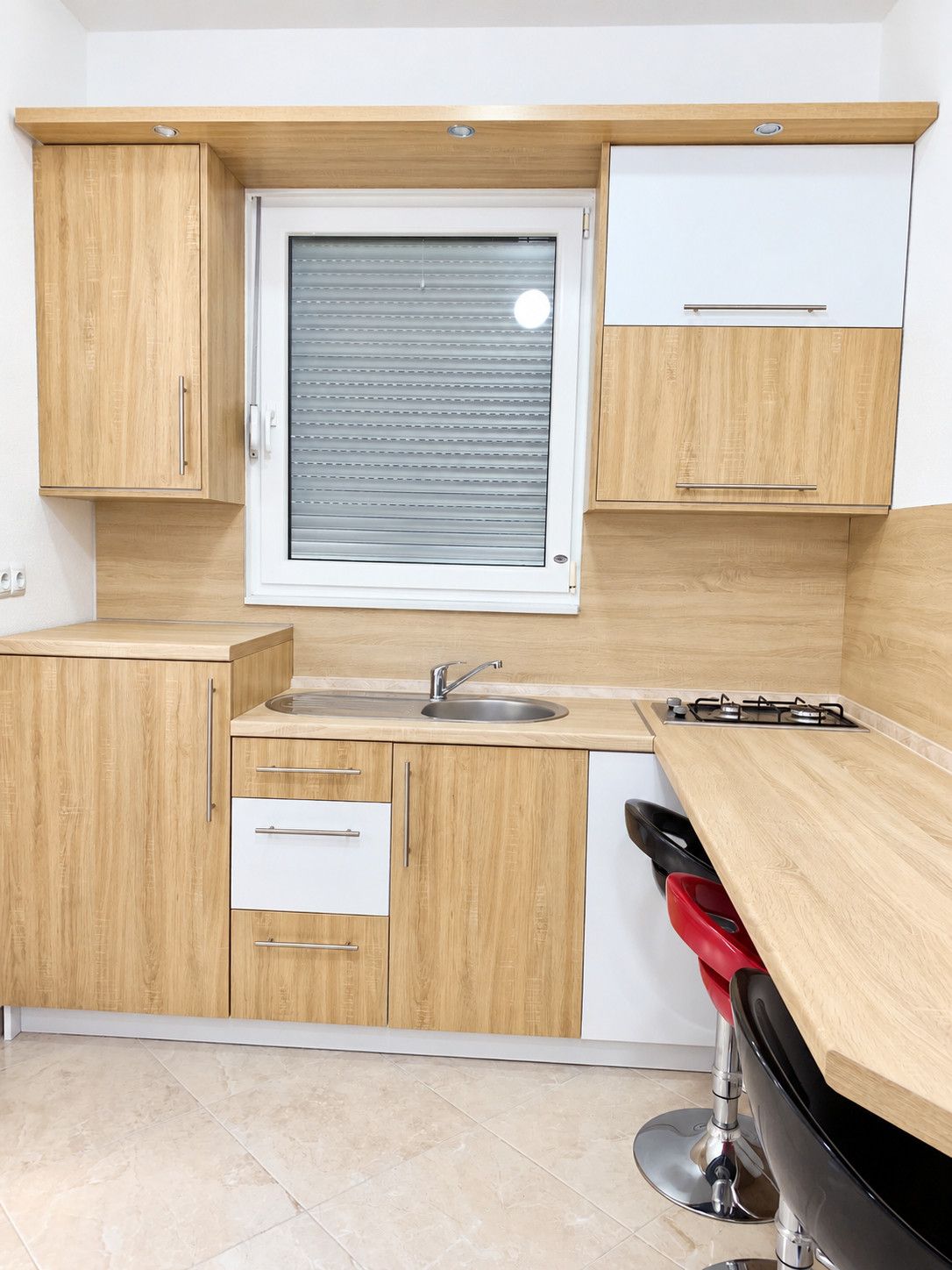 A modern kitchen with wooden cabinets, a window with blinds, a sink, a stove, and a red chair.