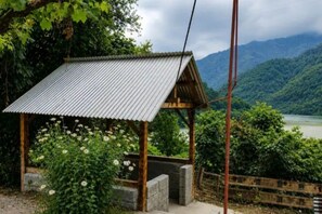 A small wooden structure with a corrugated metal roof, surrounded by lush greenery and a mountainous backdrop. - Guesthouse Noestan (Acharistskali)