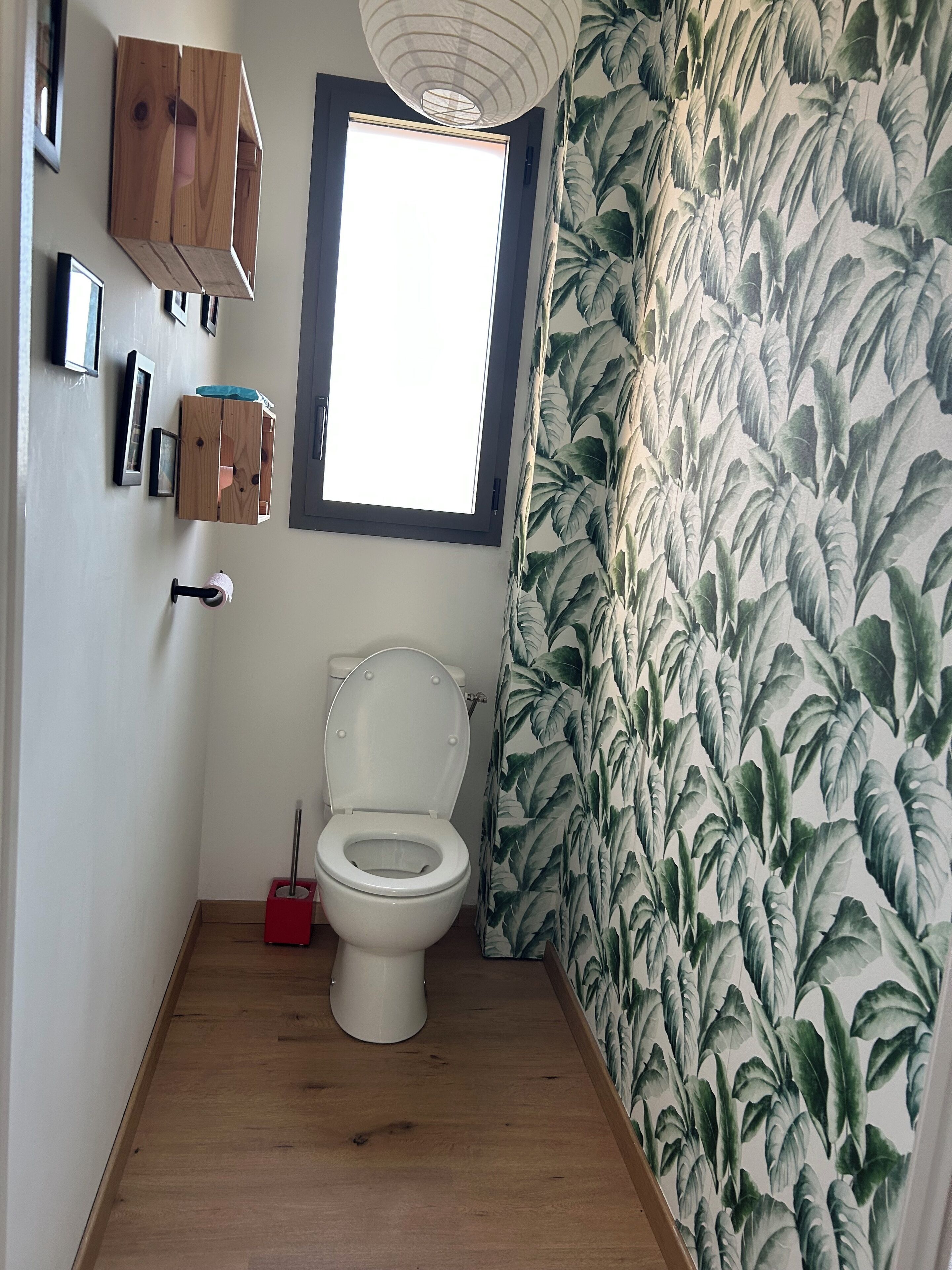 Un baño pequeño con inodoro, un armario de madera, una ventana y cortina de ducha estampada con hojas.
