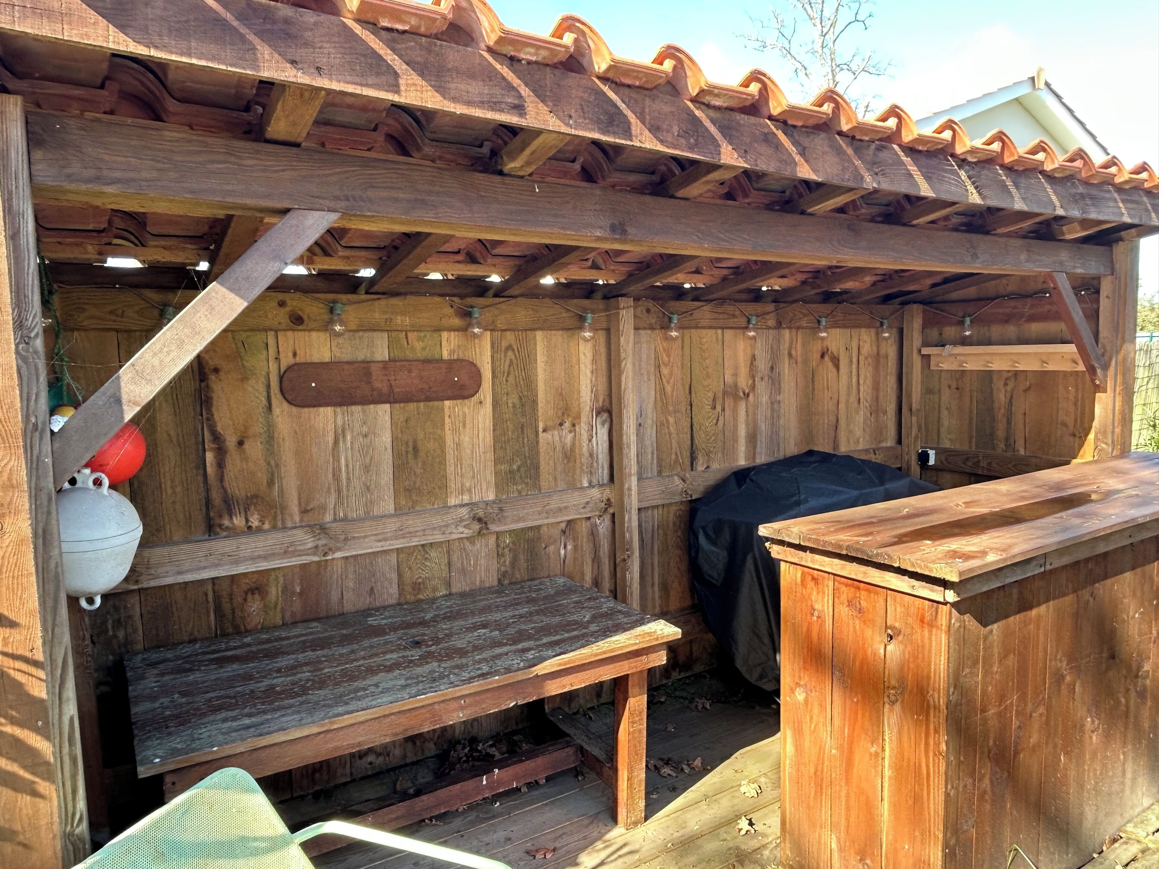 Una construcción rústica de madera con una banca, una mesa y un área cubierta con techo de tejas.