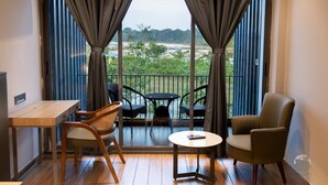 A room with a wooden floor, a small table, two chairs, and a sofa. A large window with curtains offers a view of greenery and a distant landscape. - COUNTRYSIDE RESORT MURTI (Mal)