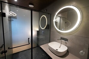 Egy modern fürőszoba üveg válaszfallal ellátott zuhanyzóval, világítással rendelkező, kör alakú tükörrel és fehér mosdókagylóval.