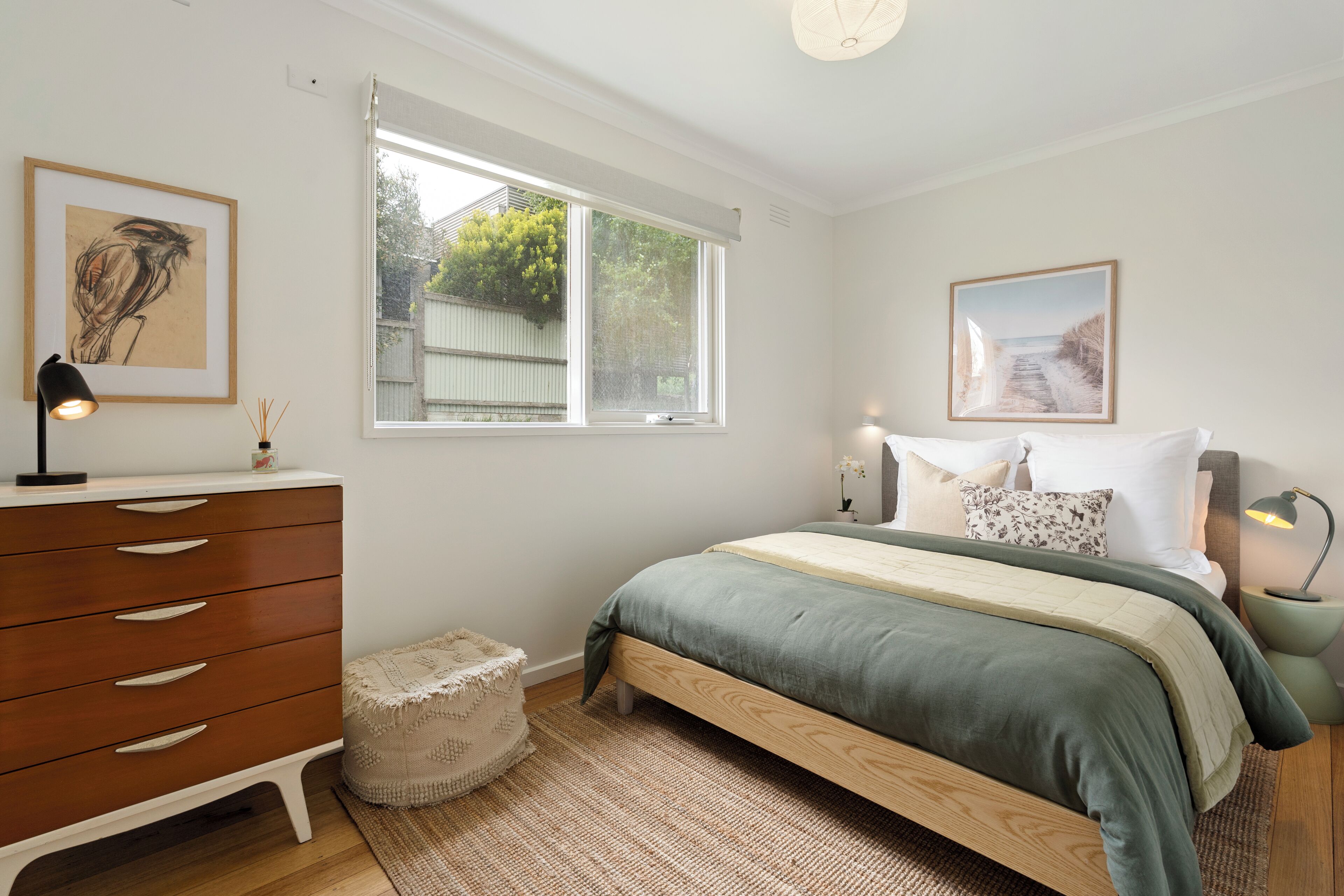 Une chambre à coucher comprenant un lit, des tables de chevet, une commode, une fenêtre et des œuvres d’art accrochées aux murs.
