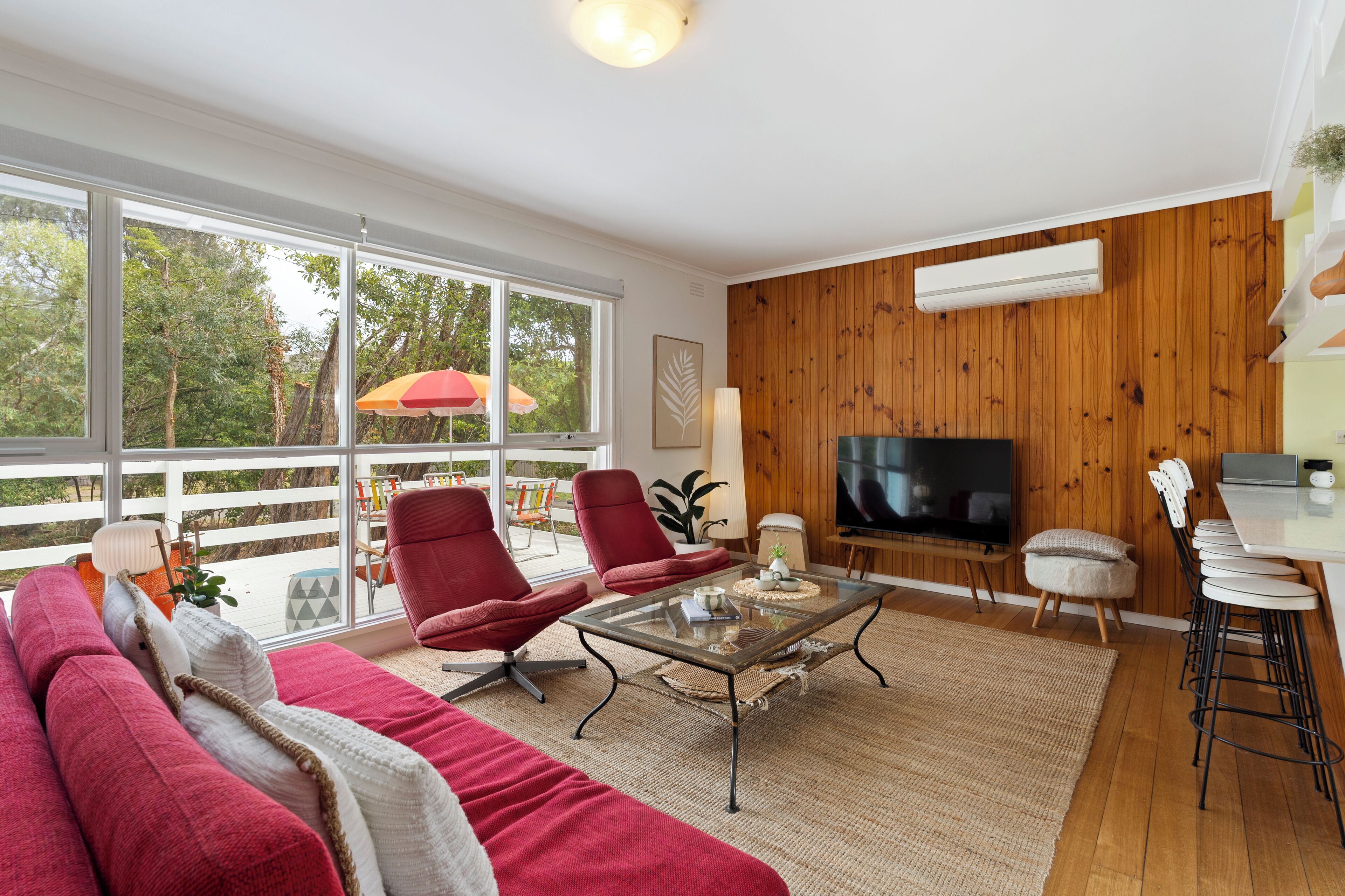 A modern living room with a large TV, red chairs, a glass coffee table, and a wooden wall.