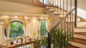 A modern interior with a curved staircase, a dining area with a round table, and a view of greenery outside. - TBK Villa Calangute (Calangute)