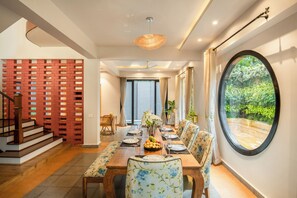 A dining area with a table set for four, a staircase, and a large window with a view of greenery. - TBK Villa Calangute (Calangute)