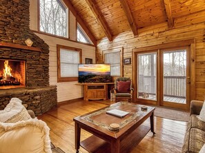 A cozy cabin interior with a stone fireplace, wooden walls, a television, and a large window. - 8 minutes from Pigeon Forge Tn/ Hot Tub/Jacuzzi Tub/ Wifi (Sevierville)