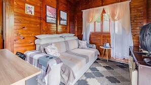 A cozy cabin interior with a wooden log wall, a sofa with striped bedding, a small table with a book, and a fan. - Casa p 5 Pessoas Perto das Praias e Beto Carrero (Penha)