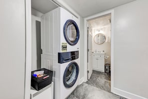 A laundry room with a front-loading washer and dryer, a black basket with items, and a small trash can. - Boutique ATL Escape Movie Room and Covered Deck (Atlanta)