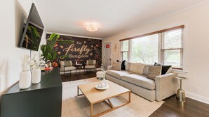 A living room with a sofa, coffee table, and a wall-mounted TV. There's a door and a window with blinds. - Boutique ATL Escape Movie Room and Covered Deck (Atlanta)