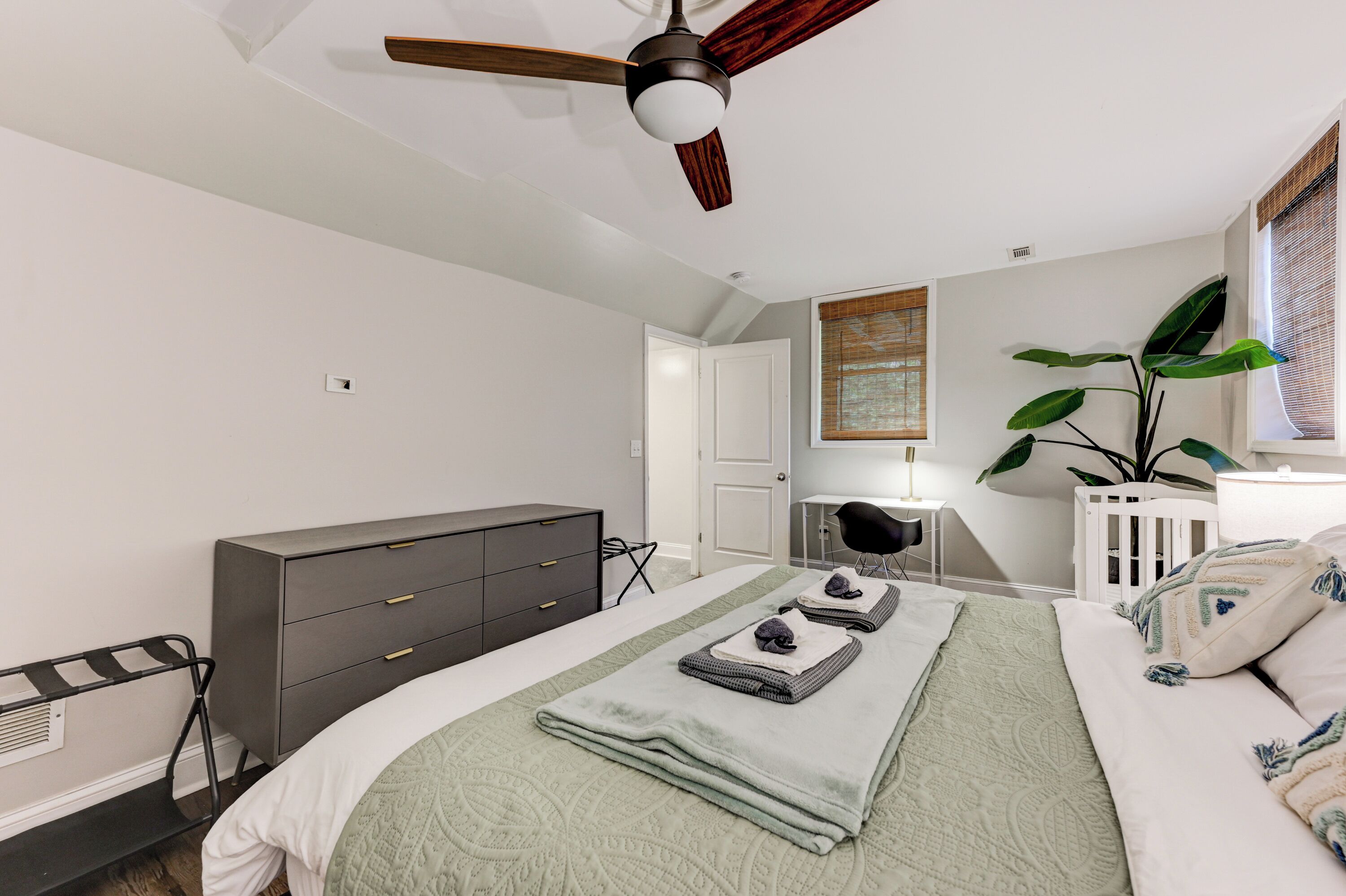 Ein Schlafzimmer mit Bett, Schminktisch, Schreibtisch, Stuhl, Deckenventilator und einer Zimmerpflanze.