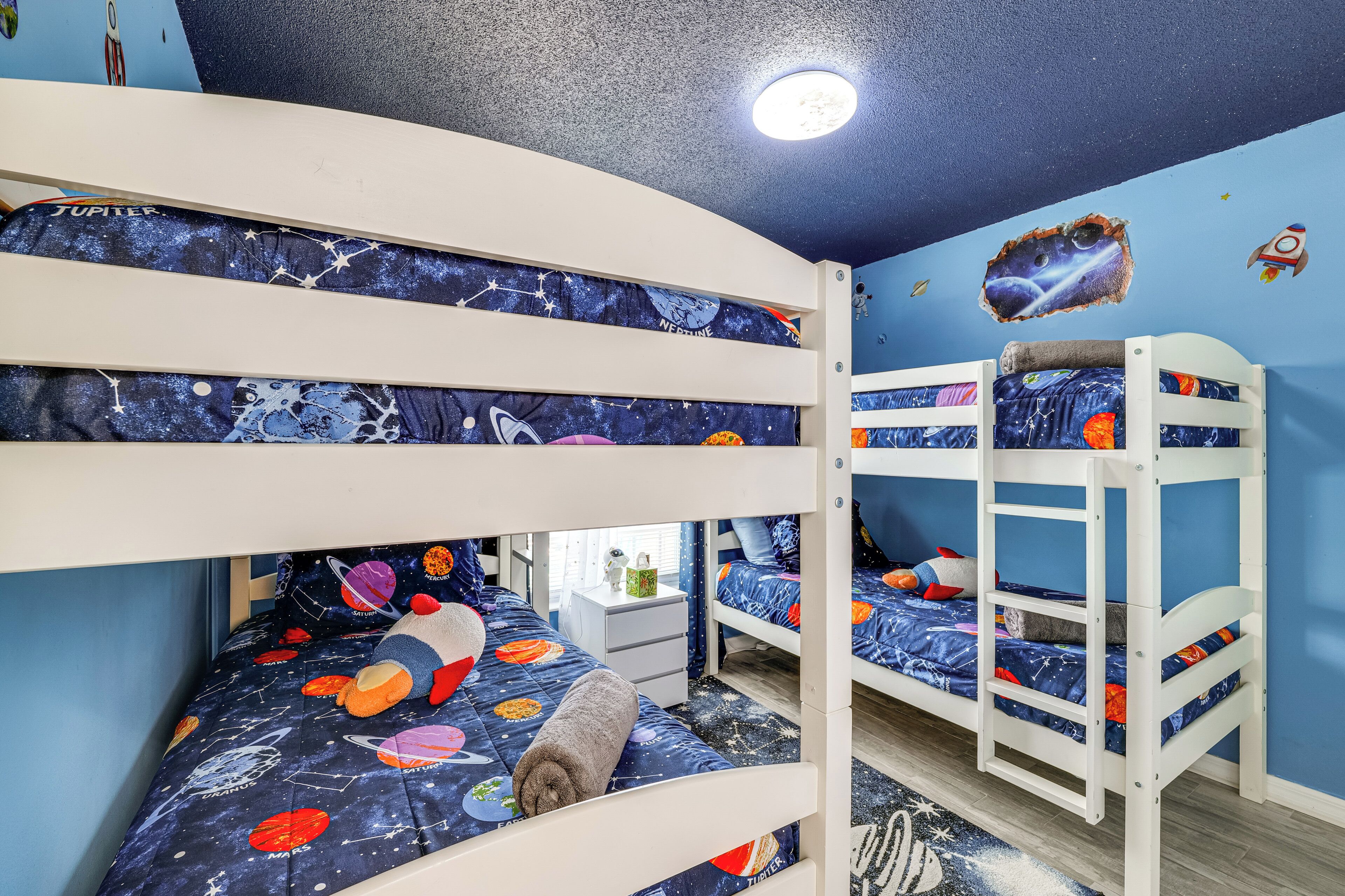 A bunk bed room with a space-themed bedspread, a small bedside table, and a wall decal of a rocket.