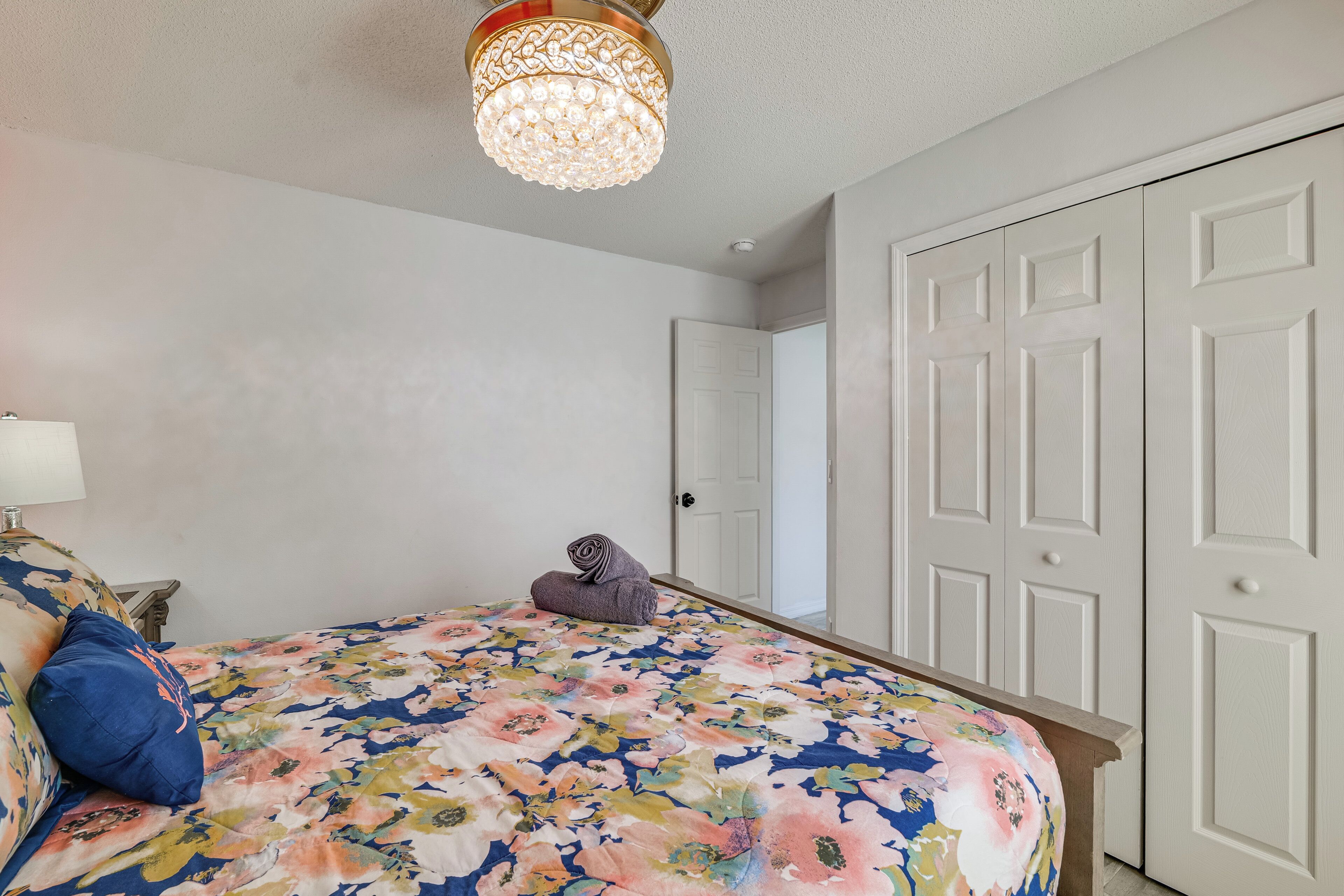 Ein Schlafzimmer mit floraler Bettdecke, einem blauen Kissen, einer dekorativen Lampe und einem Kronleuchter.