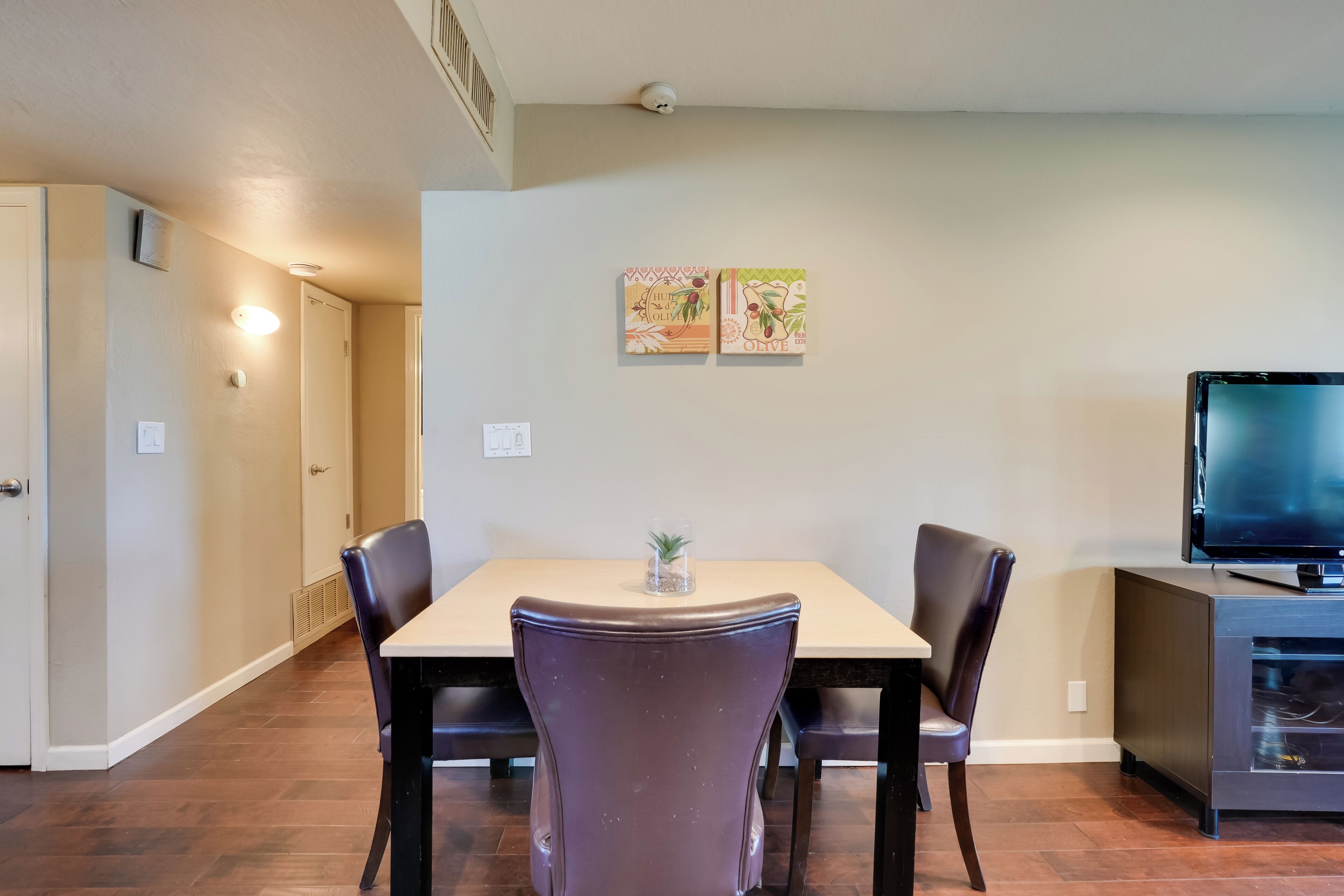 A dining area with a table and chairs, a small plant on the table, and a television on a cabinet.