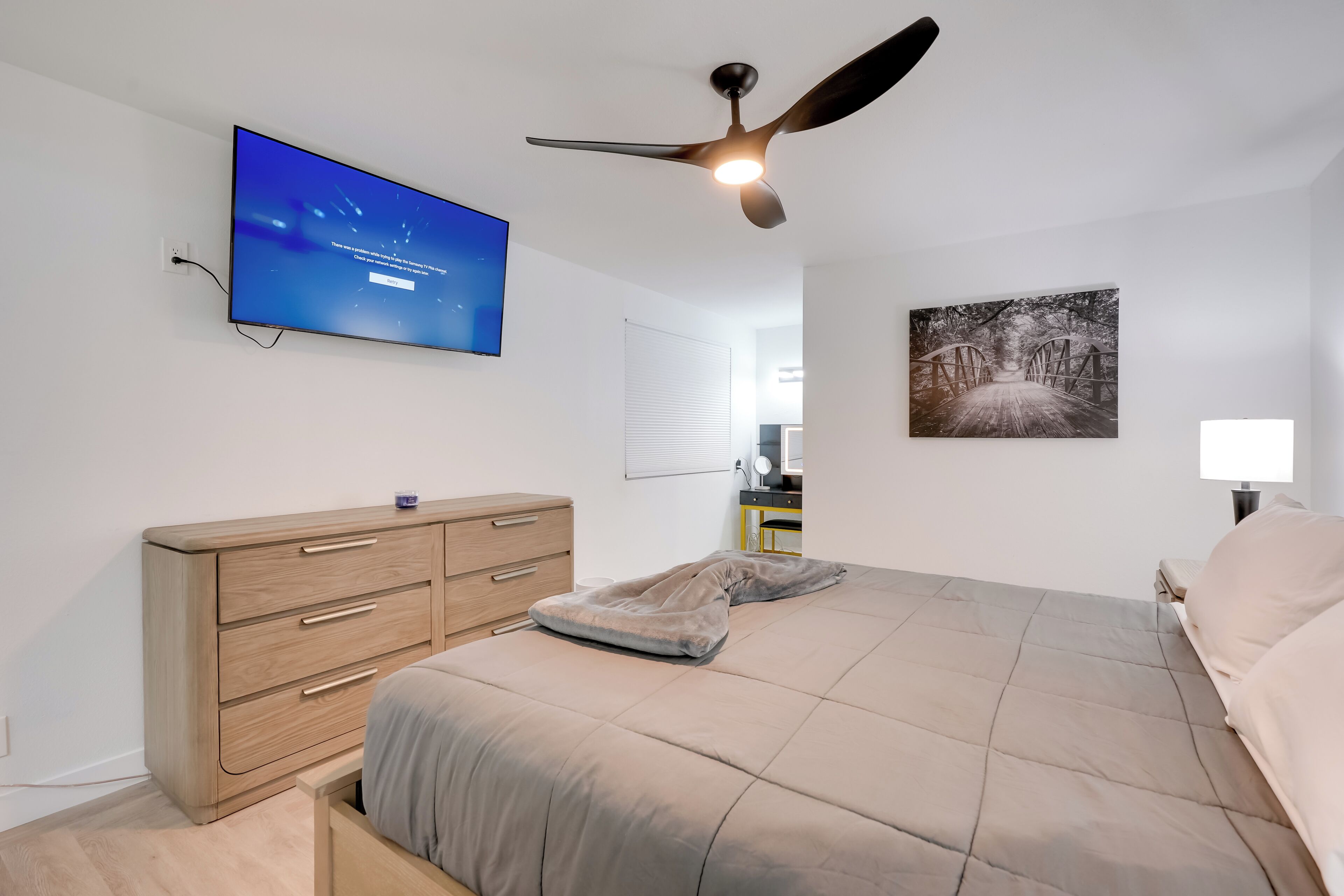 Un dormitorio con cama, ventilador de techo, televisor, cómoda y un cuadro en la pared.