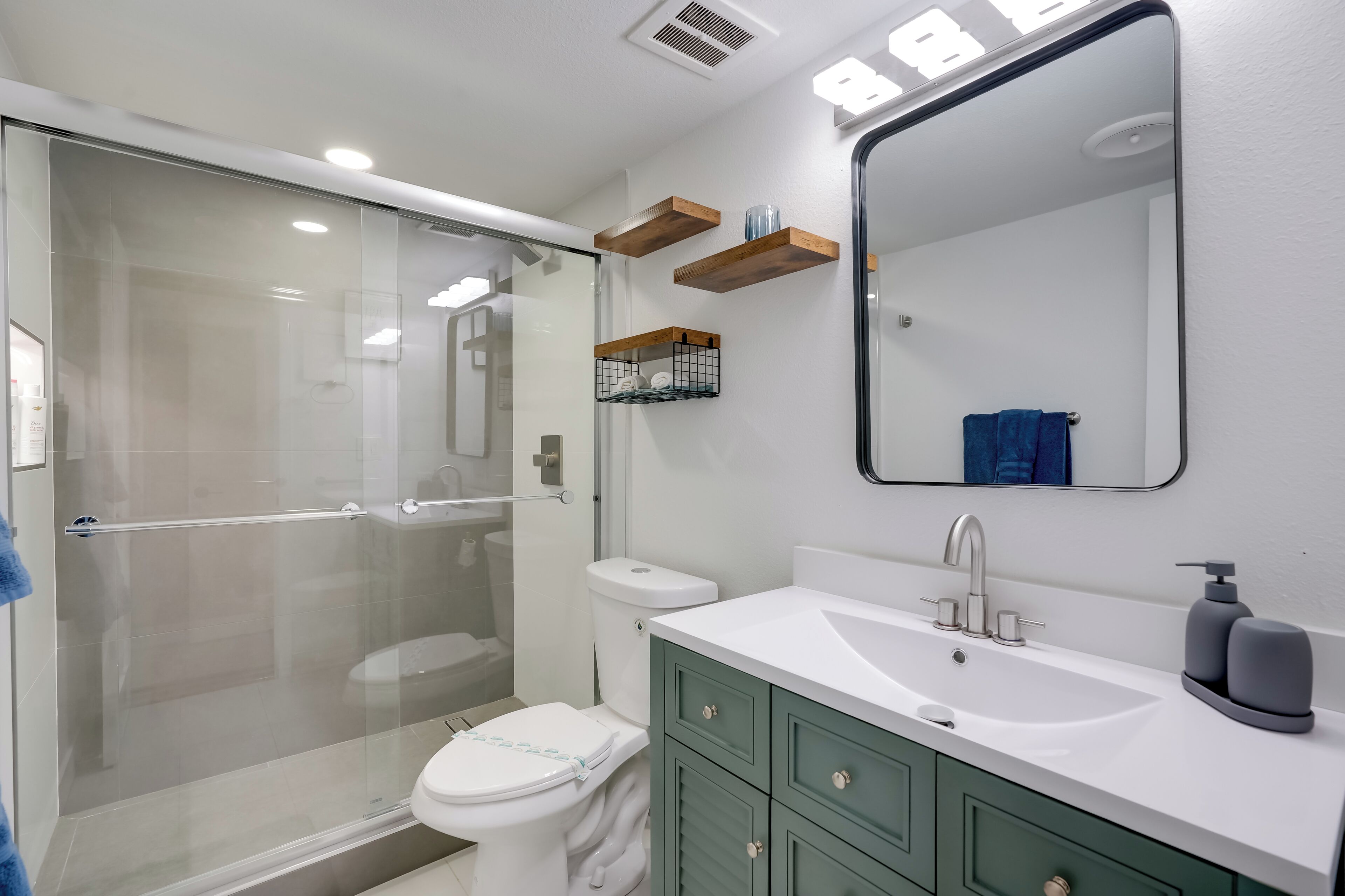A modern bathroom with a glass-enclosed shower, a white toilet, a white sink with a large mirror, and green cabinets.