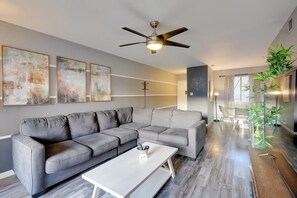 A modern living room with a grey sectional sofa, a white coffee table, and a ceiling fan. There are abstract paintings on the wall and a plant on the right. - Fully Renovated! Walkable Old Town Condo (Scottsdale)