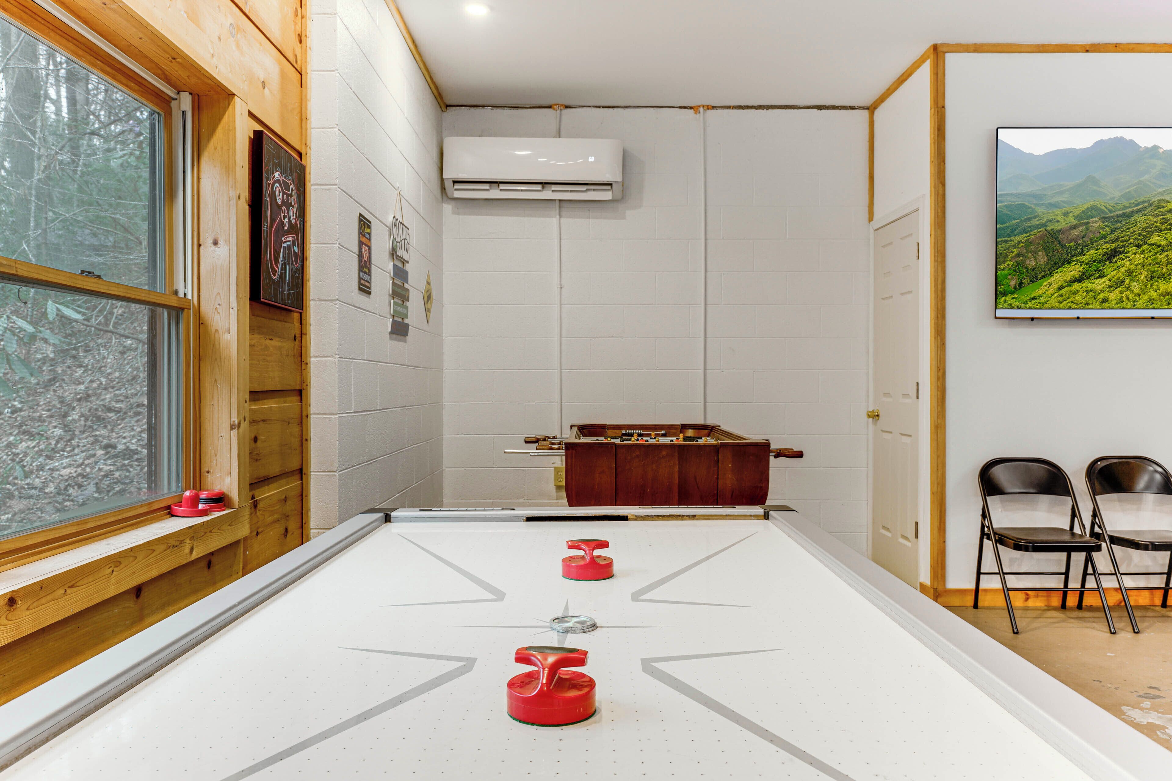 A room with a shuffleboard table, a wooden cabinet, two chairs, and a painting of a landscape.