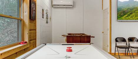 A room with a shuffleboard table, a wooden cabinet, two chairs, and a painting of a landscape.